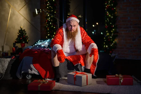 Zlý zlý Santa Claus s červeným vousem sedí na posteli a sbírá dárky na Štědrý den. Muž v obleku Santa Clause před Novým rokem. — Stock fotografie