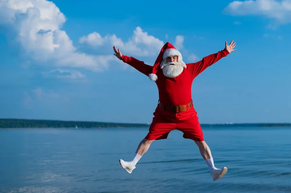 Ο Άγιος Βασίλης με σορτς πηδάει στην παραλία. Έννοια του εορτασμού των Χριστουγέννων στη θάλασσα — Φωτογραφία Αρχείου