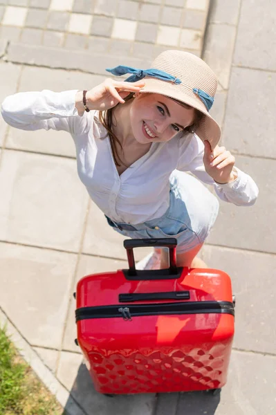 Top uitzicht op mooie jonge Kaukasische vrouw in de zomer hoed poseren met rode koffer buiten. Gelukkig lachend meisje gaat op vakantie met een grote bagage tas. Charmante dame in korte broek op een hete zonnige dag. — Stockfoto