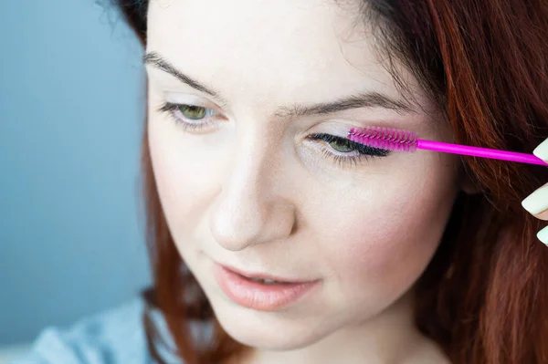 Mujer pelirroja cepillándose las pestañas de extensión con un cepillo. chica con máscara de maquillaje de párpados inminente. Industria de belleza. — Foto de Stock