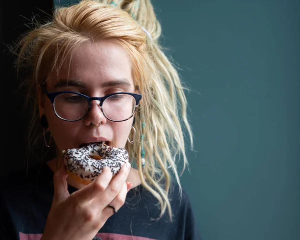 Neobvyklá dívka s dredy na hlavě a piercing jí koblihu v kavárně. Mladá žena v brýlích snídá u šedé zdi. Jíst návyky. Inzulín. — Stock fotografie