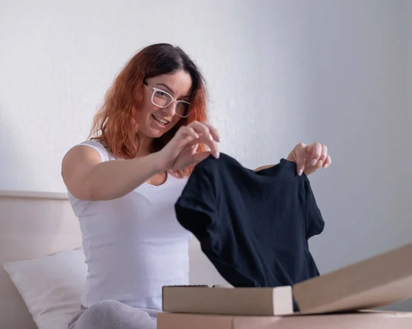 Mujer desempaqueta compra en línea de una camiseta mientras está sentado en la cama. Entrega a domicilio — Foto de Stock