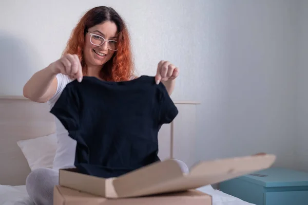 Kadın yatakta otururken internetten tişört satın alıyor. Eve teslim — Stok fotoğraf