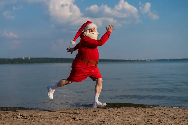 Święty Mikołaj w szortach biegnie wzdłuż plaży. Koncepcja świętowania Bożego Narodzenia na morzu — Zdjęcie stockowe