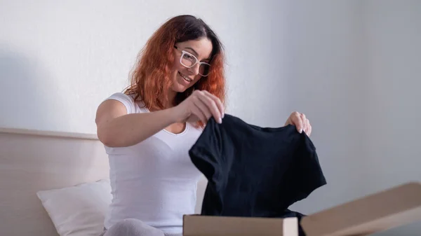 Mujer desempaqueta compra en línea de una camiseta mientras está sentado en la cama. Entrega a domicilio — Foto de Stock