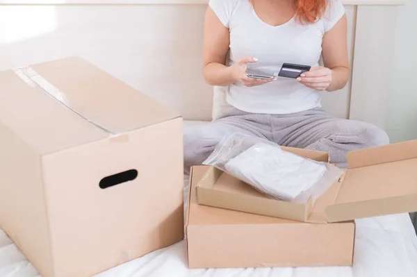 Online shopping using the application on the phone. A faceless woman sits in bed unpacks an order and holds a smartphone and a credit card. — Stock Photo, Image