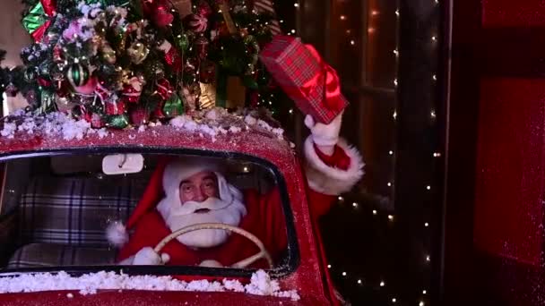 Santa Claus entrega regalos en un coche rojo y ondea una caja de regalo desde la ventana — Vídeos de Stock