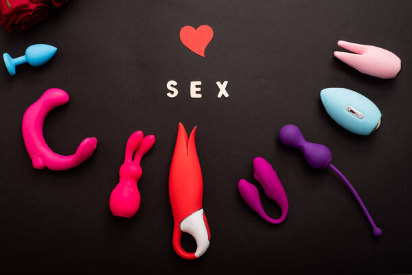 The inscription sex is a heart of roses and a collection of vibrators on a black background. Sex toys of various shapes. Assortment of sex shop