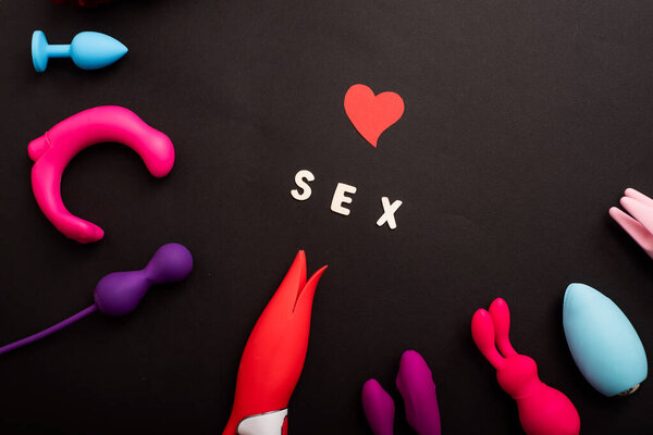 The inscription sex heart and a collection of multicolored vibrators on a black background. Sex toys of various shapes. Assortment of sex shop