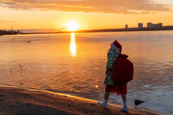 Santa claus i shorts och en skjorta går längs stranden vid solnedgången. Jul i ett varmt land — Stockfoto