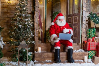 Noel Baba evin verandasında oturuyor ve bir dizüstü bilgisayar kullanıyor. Noel konsepti