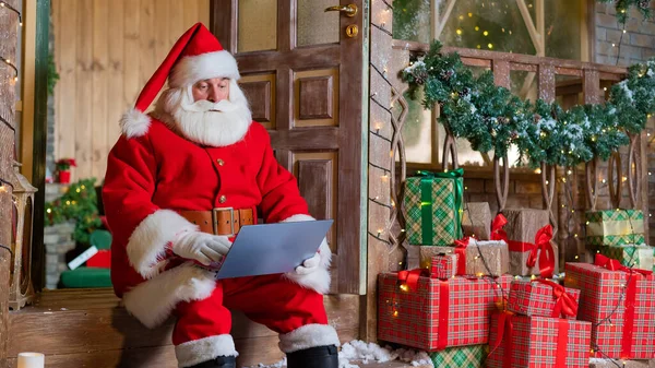 Santa Claus está sentado en el porche de la casa y utiliza un ordenador portátil. Concepto de Navidad — Foto de Stock
