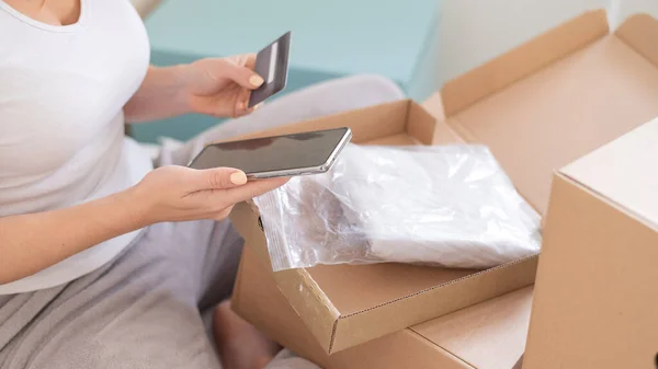 Online shopping using the application on the phone. A faceless woman sits in bed unpacks an order and holds a smartphone and a credit card. — Stock Photo, Image