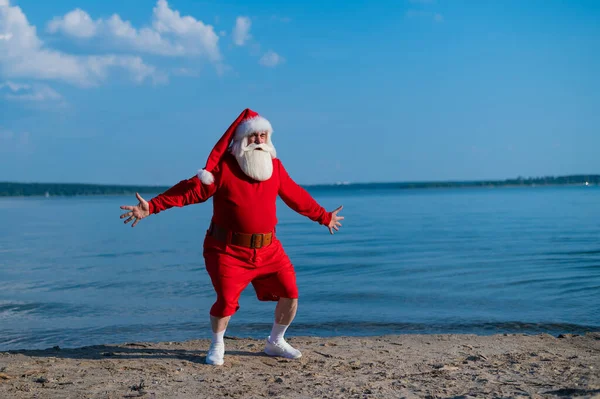 Święty Mikołaj w szortach biegnie wzdłuż plaży. Koncepcja świętowania Bożego Narodzenia na morzu — Zdjęcie stockowe
