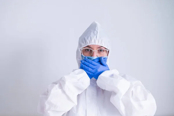 Eine Frau in Schutzanzug und Maske verhüllt ihren Mund mit ihren Händen in Handschuhen vor weißem Hintergrund. Ich spreche nicht mit dem Bösen. — Stockfoto