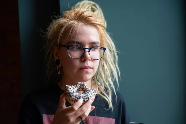 En ovanlig flicka med dreadlocks på huvudet och en piercing äter en munk på ett café. En ung kvinna i glasögon äter frukost mot en grå vägg. Ätvanor. Insulin. — Stockfoto