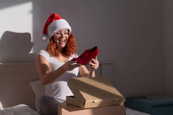 一个戴着圣诞老人帽的兴奋的女人从纸板箱里拿出红鞋子。这个女孩打开了圣诞节递送的定单。在家网上购物 — 图库照片