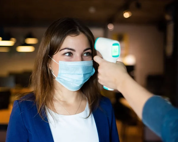 Médico mede a temperatura corporal de uma jovem mulher em uma máscara protetora com um termômetro infravermelho. — Fotografia de Stock