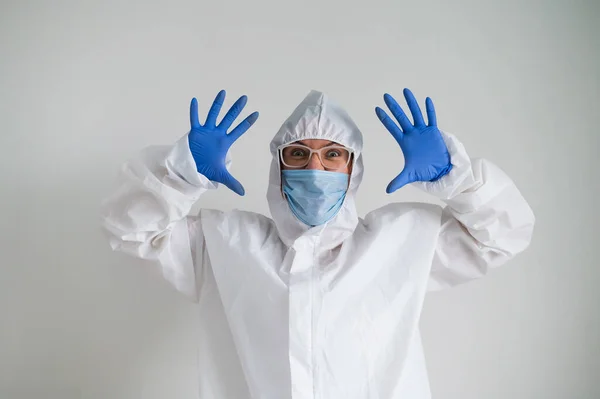 Una mujer con un traje protector y una máscara médica con una terrible mueca. El médico sostiene sus palmas a la cara y asusta — Foto de Stock