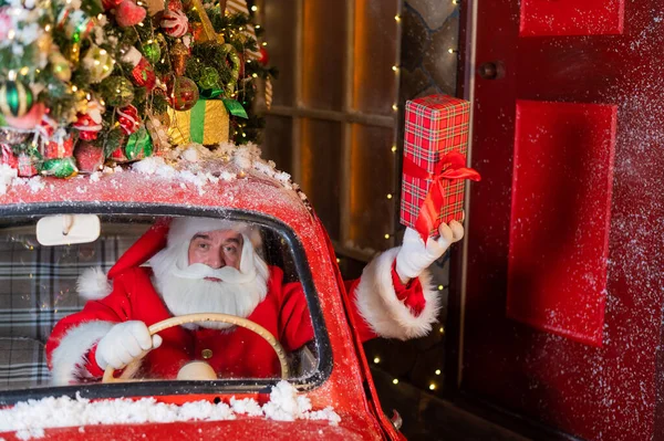 Papai Noel entrega presentes ao volante de um carro vermelho e acena uma caixa de presente pela janela — Fotografia de Stock