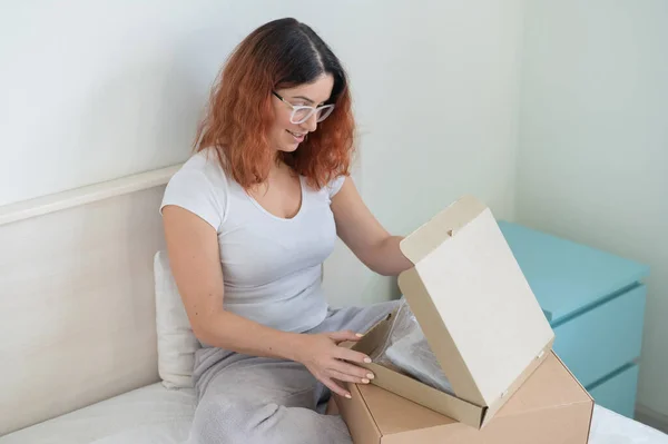 Caucasian woman opens an order while sitting in bed. Online shopping concept with home delivery — Stock Photo, Image