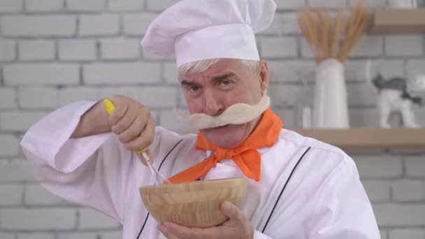 Un chef âgé fouette des œufs dans un bol en bois. Vieil homme travaillant dans la cuisine. — Video