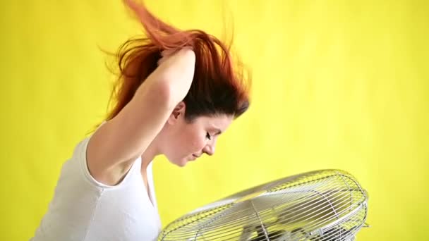 Una hermosa mujer pelirroja se enfría de pie sobre un gran ventilador eléctrico sobre un fondo amarillo. Chica con el pelo en desarrollo en el viento sobre el dispositivo para enfriar el aire. Movimiento lento. — Vídeo de stock