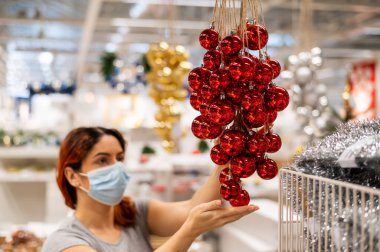 Maskeli bir kadın mağazadaki bir Noel ağacı için süsleme toplarını seçer.