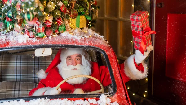 Papai Noel entrega presentes ao volante de um carro vermelho e acena uma caixa de presente pela janela — Fotografia de Stock