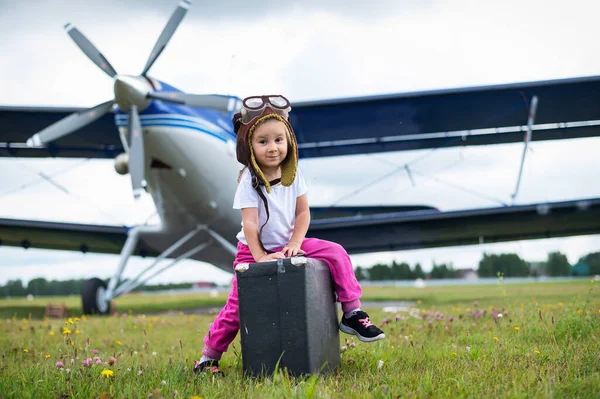 Маленькая девочка в костюме пилота носит ретро-чемодан и ходит по аэродрому. Ребенок в шляпе и очках летит на самолете. — стоковое фото