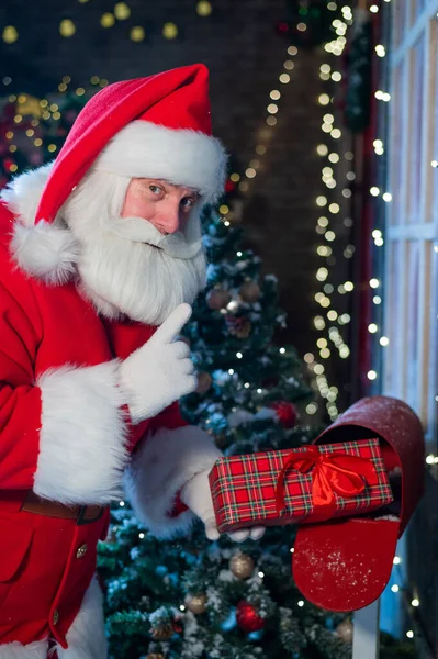 Der Weihnachtsmann verschickt ein Geschenk per Post. Ein als Weihnachtsmann verkleideter Mann legt einen geschmückten Kasten in einen Briefkasten — Stockfoto