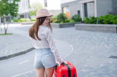 Şapkalı ve şortlu genç beyaz kadın kırmızı bavuluyla sokakta duruyor..