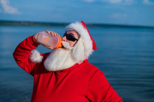Mikołaj w okularach przeciwsłonecznych pije wodę z shakera na świeżym powietrzu nad morzem — Zdjęcie stockowe