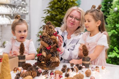 Yaşlı beyaz kadın iki torunuyla Noel için çam kozalağı süslemesi yapıyor.