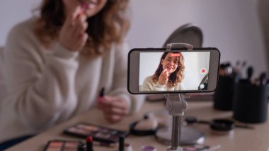 Kafkasyalı genç bir kadın cep telefonunda bir makyaj eğitimi videosu kaydediyor. Video blogcusu kız ruj sürüyor