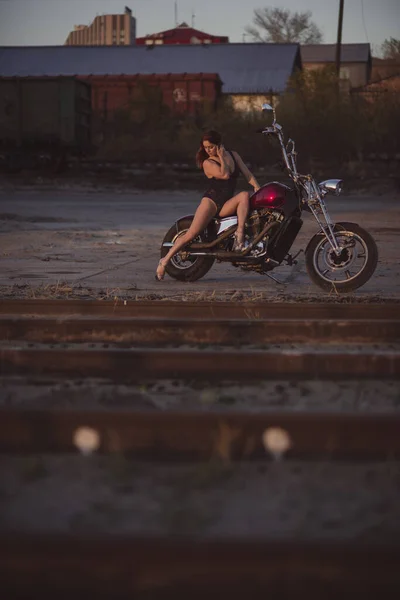 穿着花边紧身衣和高跟鞋的漂亮性感女人坐在摩托车上，背景是工业的 — 图库照片