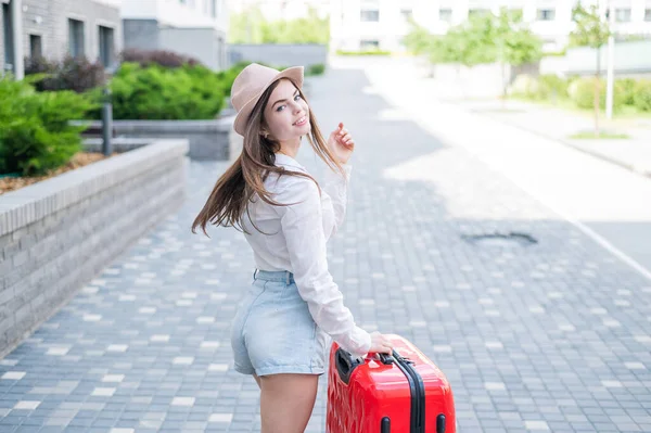 Junge Kaukasierin mit Hut und kurzer Hose steht mit rotem Koffer auf der Straße. — Stockfoto