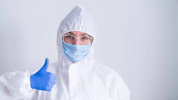 Una mujer con un traje protector y una máscara médica muestra un pulgar hacia arriba sobre un fondo blanco — Foto de Stock