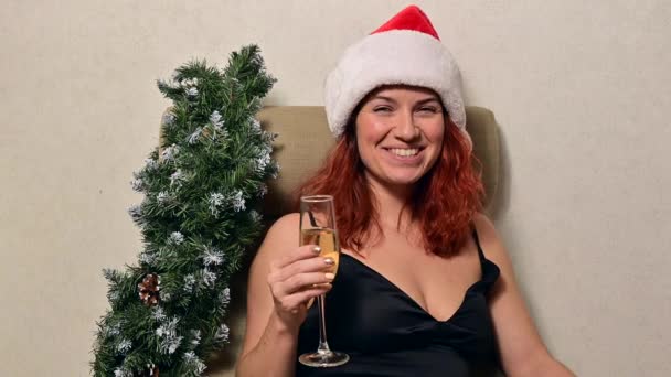 Mujer caucásica sentada en casa en un sillón con un vestido de noche y un sombrero de Santa Claus bebe champán y desea una feliz Navidad. Llamada en línea — Vídeos de Stock