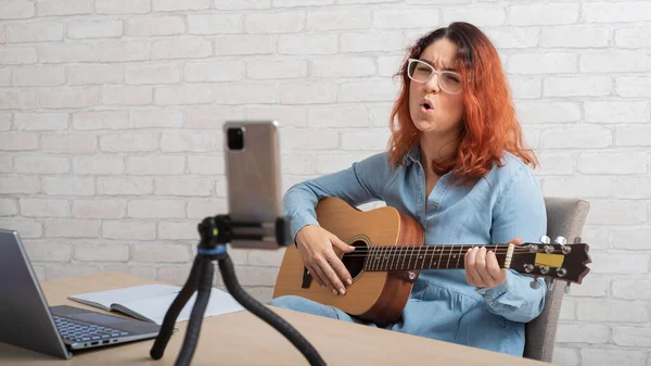 Uma mulher está transmitindo on-line em seu telefone em casa. A menina canta e toca guitarra ao vivo — Fotografia de Stock