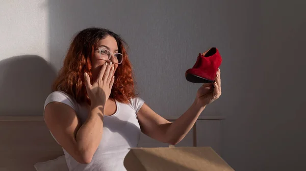 Mulher alegre abre sapatos encomendados enquanto se senta na cama. Conceito de compras on-line com entrega em domicílio — Fotografia de Stock