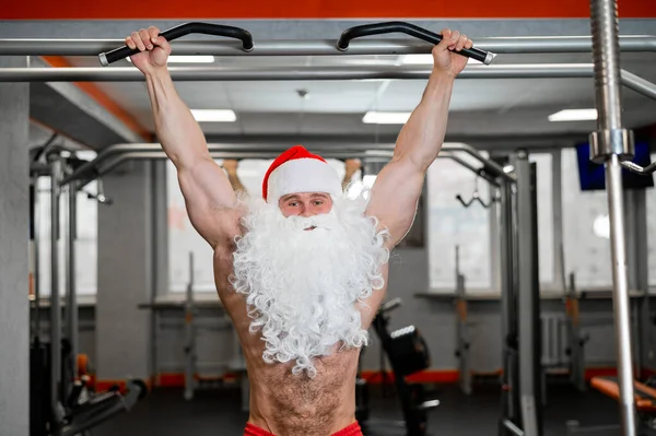 Чоловік у капелюсі Санта-Клауса і голий торс тягнеться на горизонтальну планку в спортзалі . — стокове фото