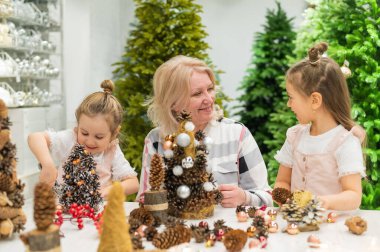 Yaşlı beyaz kadın iki torunuyla Noel için çam kozalağı süslemesi yapıyor.