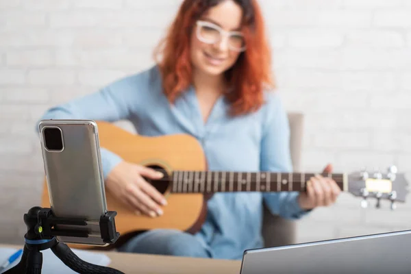 Eine attraktive rothaarige Frau bloggt über Musik. Das Mädchen singt, spielt Gitarre und nimmt Videos mit dem Smartphone auf — Stockfoto