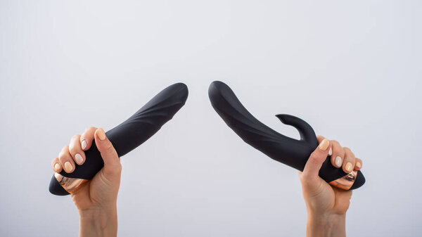 A faceless woman holds two black dildos on a white background