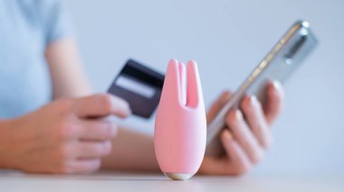 Kredi kartı akıllı telefonu olan yüzü olmayan bir kadın. Masanın üzerinde klitoral vibratör var. Çevrimiçi seks dükkanı konsepti.