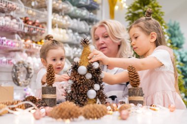 Yaşlı beyaz kadın iki torunuyla Noel için çam kozalağı süslemesi yapıyor.