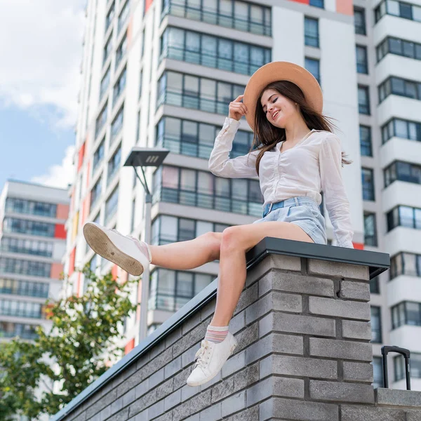 Hermosa joven en pantalones cortos y un sombrero posando mientras está sentada en un banco de piedra. Un retrato completo de una chica caucásica en un caluroso día de verano. —  Fotos de Stock