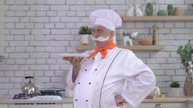 Dost canlısı yaşlı bir şef mutfakta bir tabak tutuyor. Gri saçlı, kalın gri bıyıklı bir adam mutfakta çalışıyor.