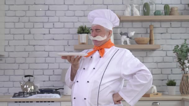 En trevlig äldre kock håller en tallrik i köket. En gråhårig man med tjock grå mustasch jobbar i köket. — Stockvideo